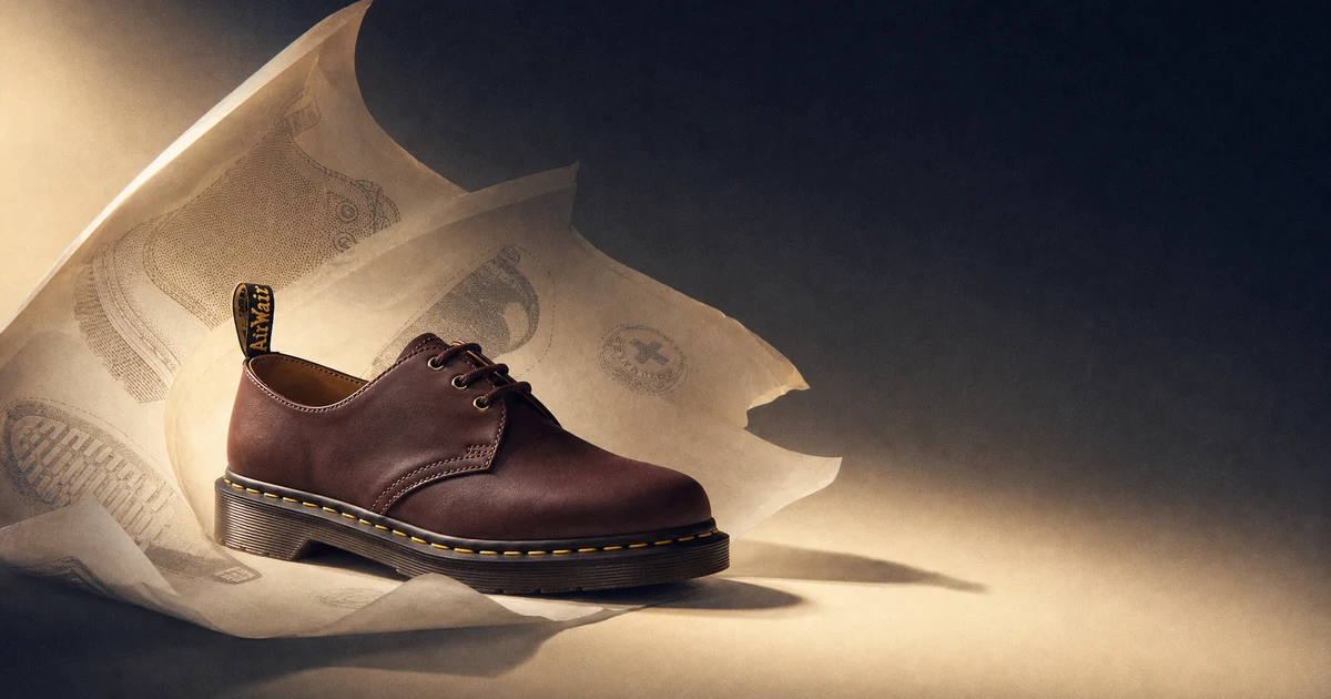 Sepatu Dr. Martens 1461 hitam dengan jahitan welt kuning dan grooved sole terlihat jelas, dikelilingi lapisan tipis seperti kertas yang terkelupas di atas latar biru gelap dengan aksen cahaya hangat